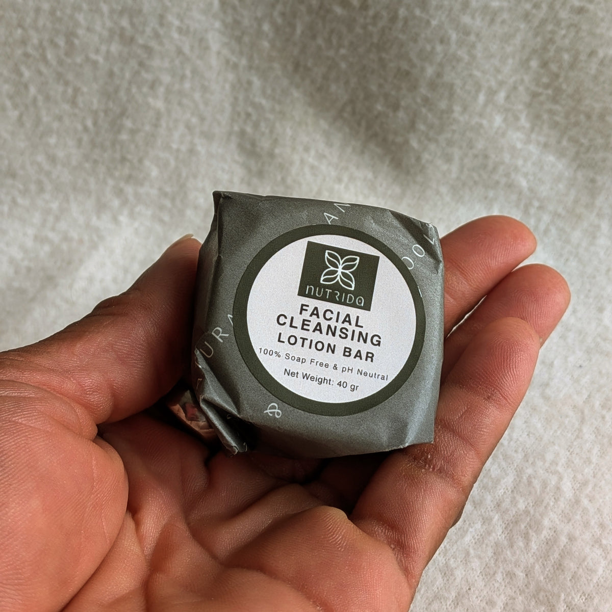 A hand holding a wrapped facial cleansing lotion bar from the brand NutriDoo, set against a soft, textured white fabric background.