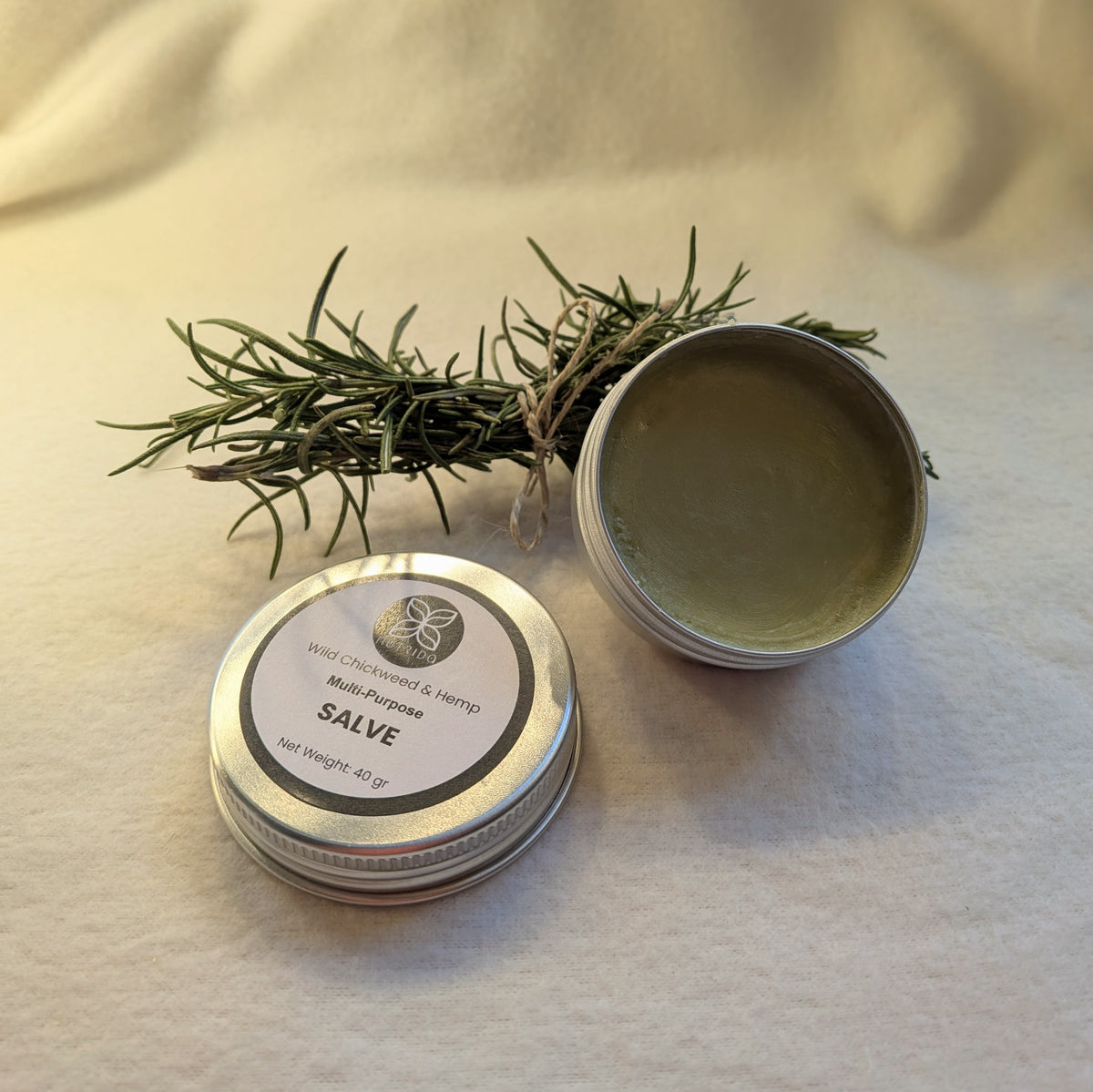 Two tins of salve with a branch of rosemary on a beige fabric background