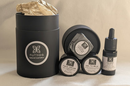 A skincare gift set from Nutrido Natural Cosmetics, featuring a large matte black cylindrical box with a gold crumpled tissue insert, three small black cosmetic jars, a small black pouch, and a black dropper bottle, all arranged on a neutral beige fabric background.