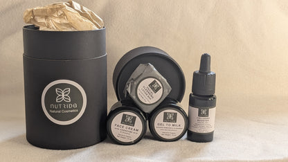 A skincare gift set from Nutrido Natural Cosmetics, featuring a large matte black cylindrical box with a gold tissue insert, alongside three smaller black containers (face cream, gel to milk, and serum) with white labels, arranged on a beige fabric background.