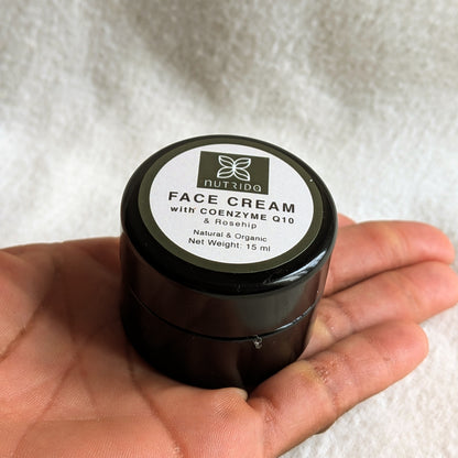 A hand holding a small black jar of NutriDQ Face Cream with Coenzyme Q10 and Rosehip, set against a soft white textured fabric background.