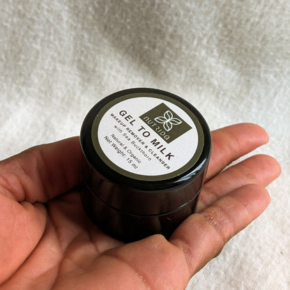 A hand holding a small black jar of Nizho Gel to Milk face cleanser against a soft white textured fabric background.