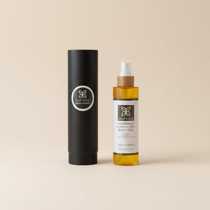 A bottle of Fuller's Calming Body Oil with a white label and amber liquid, placed next to its black cylindrical cardboard packaging on a textured white fabric background.