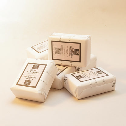 A hand holding a wrapped facial cleansing lotion bar from the brand Lotion Hero, with other wrapped bars visible in the background on a light surface.