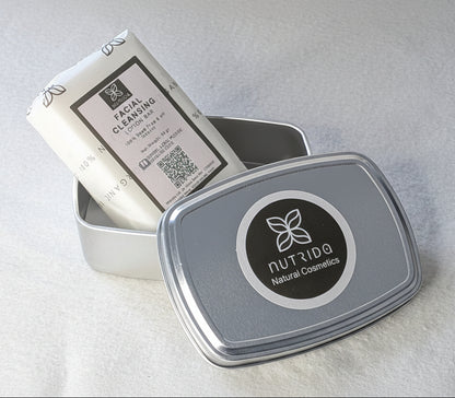 A silver metal tin with a round black-and-white logo label, shown open with a white cleansing bar product inside on a light fabric surface.