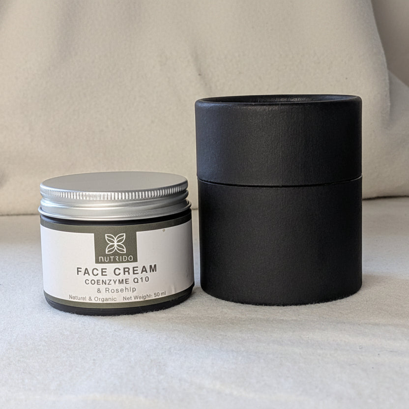 A small round jar of face cream with a silver lid and white label sits beside a tall matte black cylindrical box on a light fabric surface.