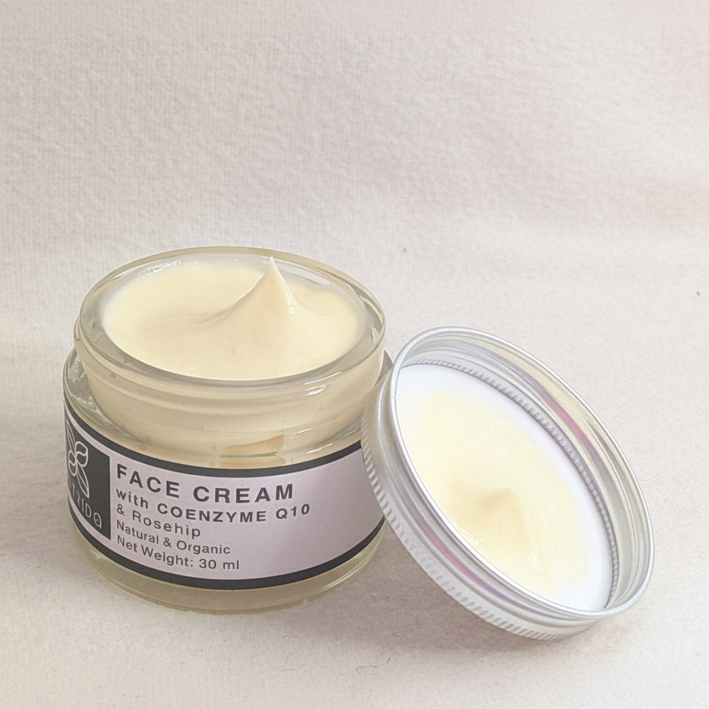 An open jar of face cream with its lid resting beside it on a light beige fabric background. The cream is a pale yellow-cream color, visible through the clear glass jar and on the lid.